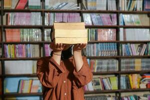 Portrait of Asian hijab woman covering her head with a pile of books in front of library bookshelf. Muslim girl reading a book. Concept of literacy and knowledge photo