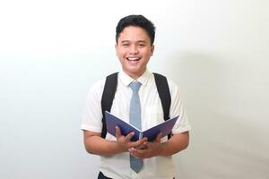 indonesio mayor alto colegio estudiante vistiendo blanco camisa uniforme con gris Corbata participación un Nota libro y sonriente mirando a cámara. aislado imagen en blanco antecedentes foto