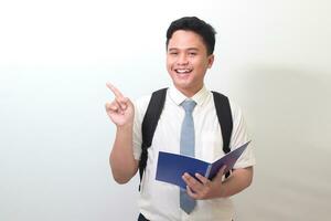 Indonesian senior high school student wearing white shirt uniform with gray tie pointing at empty space while holding an opened note book. Isolated image on white background photo