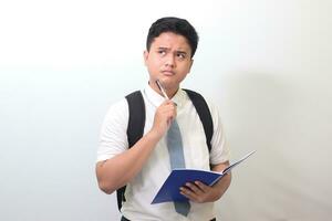 Indonesian senior high school student wearing white shirt uniform with gray tie writing on note book using pen and thinking about an idea. Isolated image on white background photo