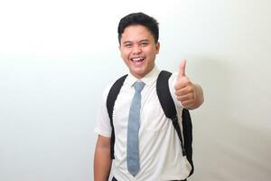 indonesio mayor alto colegio estudiante vistiendo blanco camisa uniforme con gris Corbata sonriente y mirando a cámara, haciendo pulgares arriba mano gesto. aislado imagen en blanco antecedentes foto