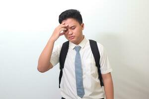 indonesio mayor alto colegio estudiante vistiendo blanco camisa uniforme con gris Corbata sufrimiento dolor de cabeza y participación manos en frente. aislado imagen en blanco antecedentes foto