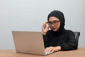 Portrait of attractive Asian hijab woman working on her laptop in office. Muslim girl adjusting glasses to see the screen clearly. Employee and freelance worker concept. photo