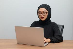 Portrait of attractive Asian hijab woman working on her laptop. Muslim girl doing task in office. Employee and freelance worker concept. photo