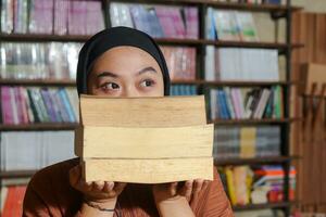 Portrait of Asian hijab woman covering her head with a pile of books in front of library bookshelf. Muslim girl reading a book. Concept of literacy and knowledge photo