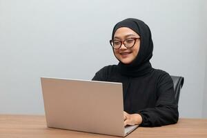 Portrait of attractive Asian hijab woman working on her laptop. Muslim girl doing task in office. Employee and freelance worker concept. photo