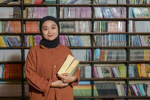 retrato de asiático hijab mujer participación libro en frente de biblioteca estante para libros. musulmán niña leyendo un libro. concepto de literatura y conocimiento foto