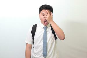 indonesio mayor alto colegio estudiante vistiendo blanco camisa uniforme con gris Corbata mirando mediante dedos, echar un vistazo en conmoción cubierta cara y ojos con mano. aislado imagen en blanco antecedentes foto