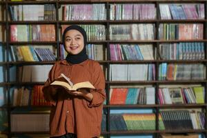 Portrait of Asian hijab woman holding book in front of library bookshelf. Muslim girl reading a book. Concept of literacy and knowledge photo