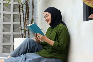 Portrait of attractive Asian hijab woman reading book in balcony. Muslim girl enjoying free time. Concept of literacy and knowledge photo