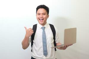 indonesio mayor alto colegio estudiante vistiendo blanco camisa uniforme con gris Corbata participación un ordenador portátil y demostración pulgar arriba mano gesto, en pie en contra gris antecedentes foto