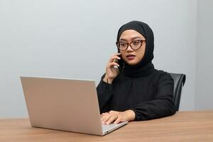 Portrait of attractive Asian hijab woman working on her laptop. Muslim girl making a phone call in office. Employee and freelance worker concept. photo