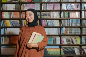 Portrait of Asian hijab woman holding book in front of library bookshelf. Muslim girl reading a book. Concept of literacy and knowledge photo