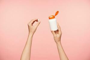 Indoor shot of bottle with vitamins being held by young fair-skinned female's hands being isolated over pink background. Healthcare and beauty concept photo