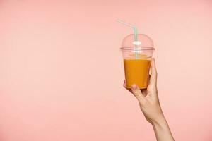 Studio photo of raised well-groomed woman's hand with nude manicure holding plastic cup of orange juice with straw while being isolated over pink background