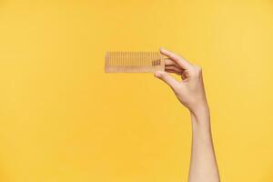 Cropped shot of young female's hand being raised while holding wooden hairbrush with forefinger and thumb being isolated against orange background photo