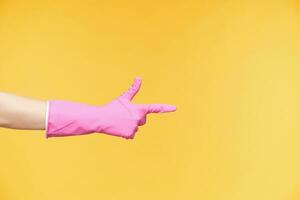 Indoor shot of raised hand in pink gloves showing index finger while pointing aside, posing over orange background. Cleaning and gesturing concept photo