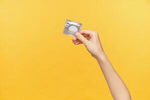 Horizontal shot of young fair-skinned female's hand being raised while holding silver pack with condom. Young woman prefer safe sex, posing over orange background photo