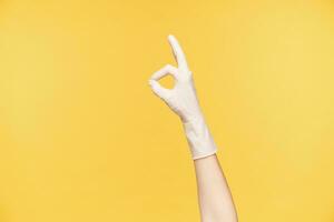 Indoor shot of raised female's hand in rubber white glove forming with fingers well done gesture, finishing spring cleaning and being satisfied, posing over orange background photo