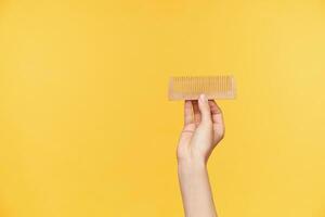 Indoor photo of young fair-skinned female's hands with nude manicure holding wooden hairbrush horizontally while being isolated over orange background