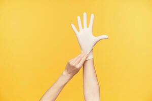 Indoor photo of young female's hand being raised while taking on rubber glove and keeping all fingers separately, isolated against orange background