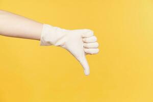 Indoor shot of fair-skinned raised hand dressed in rubber glove thumbing down while expressing negative emotions, isolated against orange background photo