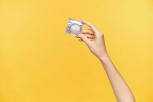 Indoor photo of young woman's raised hand with nude manicure keeping silver pack with condom while posing over orange background. Relations and sex concept