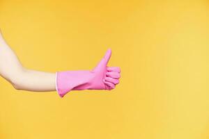 Side view of raised hand in rubber gloves thumbing up while showing positive emotions, being satisfied while finishing spring cleaning, isolated over orange background photo