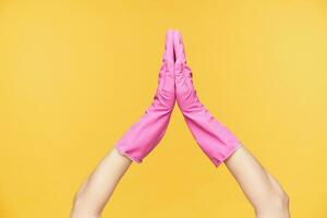 Horizontal photo of raised female's hands in rubber gloves keeping palms together while being isolated over orange background. Body language and gesturing concept