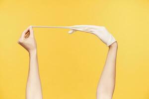 Studio photo of young woman's hands posing over orange background, one hand pulling middle finger while taking out white rubber glove. Human hands concept