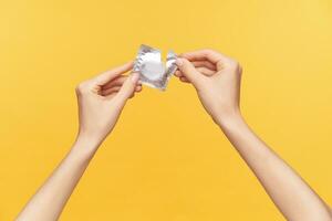 Cropped shot of young raised female's hands keeping silver pack with condom and unpacking it while being isolated over orange background. Relations and sex concept photo