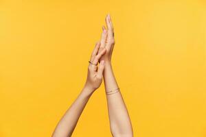 Studio photo of raised fair-skinned woman hands with nude manicure moisturizing it with cream while posing over yellow background. Bodycare concept