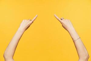 Horizontal shot of yound female hands with nude manicere keeping forefingers raised while showing with it on point in middle, isolated over yellow background photo