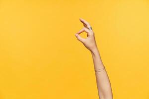 Indoor photo of well-groomed fair-skinned female hands with ornamentals being raised while showing ok gesture with fingers, isolated over yellow background