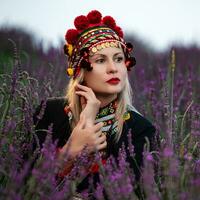 A girl in a chelsea headdress and a black dress decorated with red embroidery sits between lavender bushes. photo