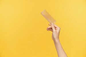 Indoor shot of young pretty woman's hand raising up wooden hairbrush while posing over orange background, young woman going out and making hairdo photo