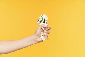 Studio photo of smashed ice-cream by fair-skinned female's hand while posing over yellow background, having diet and refusing of sweets. Food and diet concept