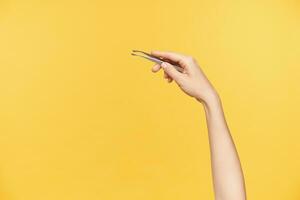 Indoor photo of young woman's hand being raised while keeping pincers in it, going to tweeze eyebrows while being isolated over orange background. Beauty and face care concept