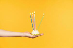 Side view og elegant lady's hand holding delicious cake with lightened colored candles while receiving bithday congratulations, isolated over yellow background photo