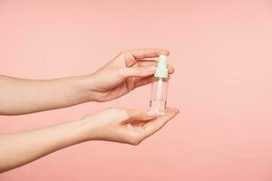 Indoor shot of raised female's hand with nude manicure holding spray bottle with disinfectant while being isolated over pink background. Human hands concept photo