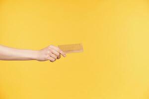 Indoor photo of pretty female's hand reaching out ahead while holding wooden comb, being isolated over orange background. Human hands and body language concept