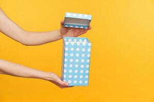 Cropped shot of young fair-skinned woman unpacking mint dotted holiday box with gift while standing over yellow background. Celebrating and events concept photo