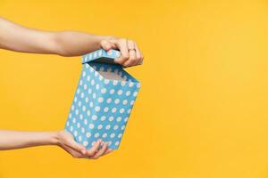 Horizonatal photo of pretty lady's hand with nude manicure geeting surprise and unpacking present mint dotted box while standing over yellow background