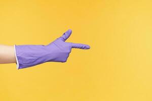 Studio photo of fair skinned hand in rubber glove being raised while pointing aside with index finger, being isolated over yellow background while cleaning house