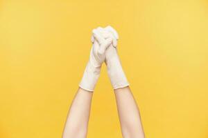 Studio shot of raised young woman's hands in rubber gloves folding together and keeping fingers crossed while being isolated over orange background photo
