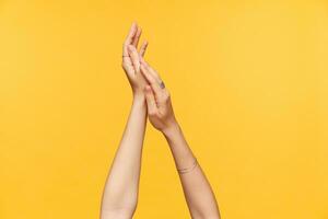 Cropped shot of well-groomed fair-skinned hands of young female being raised while putting cream on it, isolated over yellow background. Hands and body care concept photo