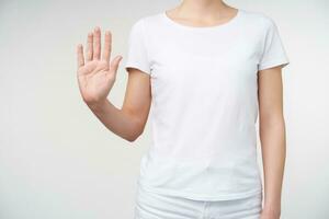 Shot of young female dressed in white t-shirt showing raised palm at camera while pointing up with fingers number five, standing over white background photo