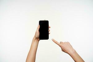 Studio shot of young woman's hands being raised while keeping smartphone in it and showing on screen with index finger, isolated over white background photo