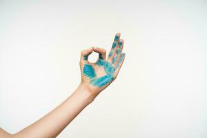 Indoor photo of young lady's arm painted with blue colour forming namaste sign while posing over white background, meditating with raised hand