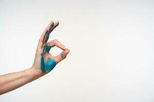 Side view of raised young female's hand painted in blue colour forming with fingers well done sign while standing over white background. Signs and gesturing concept photo